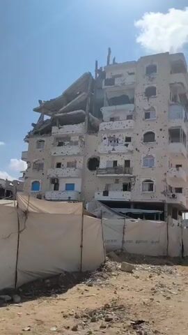 The video shows a severely damaged multi-story building. The top floors of the building appear to be partially collapsed, with exposed rebar and broken concrete. The exterior walls are heavily pockmarked with what appear to be bullet or shrapnel holes. Several windows are broken or missing, and some balconies are damaged. In the foreground, there is a temporary fence or barrier, possibly made of fabric or tarpaulin, with some visible writing that includes the word "UNICEF". The ground in front of the building is sandy and appears to be littered with debris. The sky is visible and appears to be clear. No people are clearly visible in the video. The overall scene depicts a structure that has been subjected to significant destructive forces.