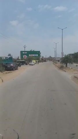 The video shows a mostly empty two-lane road, seen from the perspective of someone moving along it. The sky is light blue with scattered clouds. A green building with "Power Shield" written on it is visible on the left side of the road. There are parked cars and motorbikes near the building. A couple of people are walking along the side of the road. Further down the road, there are a few vehicles, including a car and an auto rickshaw. Light poles line the road. The shoulder of the road is unpaved and sandy.