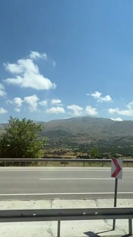 A handheld video shows a view from inside a vehicle looking out at a roadside scene with a mountain range in the distance. The sky is blue with scattered white clouds. A paved road runs horizontally across the lower portion of the frame, with a white line marking the lane. A guardrail is visible in the foreground. Beyond the road, there is a dry, grassy area with scattered trees and shrubs. A road sign with a red and white chevron pattern is visible on the left side of the road. The background is dominated by rolling hills and mountains. The video is shaky, indicating movement.
