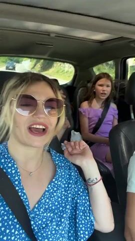 The video shows a family in a car, with three young girls in the back seat. The girl in the front left is wearing a blue and white patterned top and sunglasses. The girl in the middle back is wearing a green and white top with butterfly patterns. The girl on the right back is wearing a purple off-the-shoulder dress. They are all wearing seatbelts and appear to be on a road trip.

The scene is set inside a car, with black leather seats and a sunroof. Through the windows, we can see a sunny day with lush green hills and a road with other cars. The girls seem to be engaged in conversation or singing, with expressions of excitement and enjoyment. The girl in the middle back is gesturing with her hands, and the girl on the right back is looking towards the camera and speaking.

There are a few objects visible in the back seat, including a bottle of water, a dark colored piece of clothing, and a small brown object, possibly food. There is no visible text in the video.

The overall mood is cheerful and lively, suggesting a fun family outing. The sunlight filtering in suggests it is daytime. The scenery of rolling green hills hints at a potentially rural or scenic location, which is consistent with the provided context of Brajici, Montenegro.
