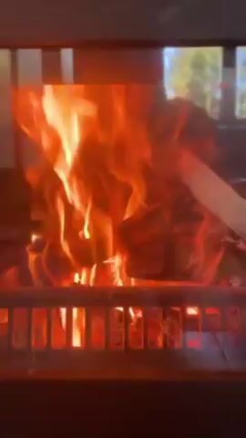 The video depicts a close-up shot of a blazing fire within a fireplace. Flames lick upwards and outwards, visible through the metal grate supporting the burning wood. The light from the fire illuminates the interior of the fireplace and casts a warm glow. The environment outside the fireplace is slightly visible but blurred and indistinct.