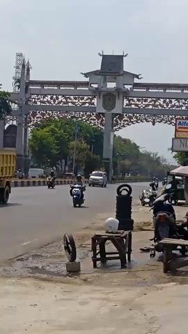 The video shows a roadside scene in what appears to be a city in Indonesia. In the foreground, there is a tire repair stand with several tires stacked vertically and a white helmet placed on a small wooden stool. A person, likely the vendor, is sitting on a wooden bench to the right. In the middle ground, a two-lane road is visible with several motorcycles and a white car driving. On the left side of the road, there are trees and buildings. Dominating the background is a large, ornate archway, possibly a gateway to a city or district, featuring intricate carvings and traditional architecture. The sky is bright and clear, indicating a sunny day. The overall impression is of a busy urban environment with street-level commerce.