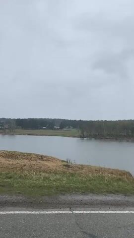 The video shows a view of a lake on a cloudy day, shot from a moving vehicle.  The lake is calm and gray under an overcast sky.  Along the near shore, there is a strip of grass and a road.  Across the lake is a treeline and some buildings in the distance.  The camera is moving along the road, providing a passing view of the lake.