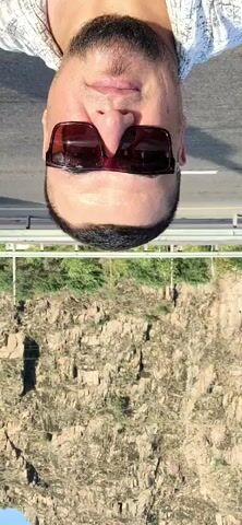The video begins with an extreme close-up shot of a man's face, upside down. He is wearing dark sunglasses with thick frames and has a beard. The man appears to be smiling slightly. The background is out of focus, but it seems to be a road with a white vehicle on the right side of the frame. As the video progresses, the camera slowly pans down, revealing a mountainous landscape with rocky cliffs and sparse vegetation. The sky is a clear blue. The man's face remains the central focus of the video. The video is short, lasting only a few seconds, and there is no audible sound.
