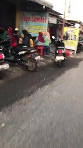 The media file shows a street scene in Balonggabus, Indonesia. It appears to be daytime, with overhead lighting suggesting it could be late afternoon or early evening. The ground is wet, indicating recent rain or the presence of puddles, and the lighting is somewhat muted, possibly due to overcast skies or the time of day.

The primary subjects are several people and a few motorbikes. A group of individuals is gathered near a building with a prominent sign. One person is seated on a red stool, while others are standing or leaning. Their clothing suggests a casual setting, with some individuals wearing headscarves. The motorbikes, parked on the street, are a common mode of transportation in the area.

The building in the background has a colorful sign with Indonesian text. The visible text reads "Sago Sambel 10 Macam." This suggests that the establishment is likely a food stall or restaurant selling sago dishes and sambal (chili paste) with ten different varieties. Another yellow sign is partially visible to the right, but the text is illegible.

The overall atmosphere seems calm, with a sense of everyday activity. There are no overt signs of distress or urgency. The scene depicts a typical street in a local Indonesian community, likely a commercial area with shops or eateries. The blurred motion of the video suggests it might have been recorded while moving, possibly from a passing vehicle or while walking.