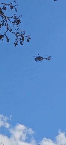 A helicopter is flying in a clear blue sky with some puffy white clouds. A bare tree branch is in the upper left corner of the frame. The helicopter is small in the frame and appears to be moving slowly.