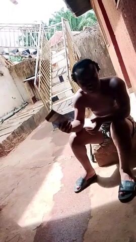 A shirtless man sits on a ledge in an outdoor area next to a building and stares at a device in his hands. He is wearing dark-colored shorts and green and black sandals. To his left is a set of stairs made of metal and concrete. The area around him is paved with a dirt-colored material. There is a building next to him with a red wall. Other buildings are visible in the background. Music can be heard.
