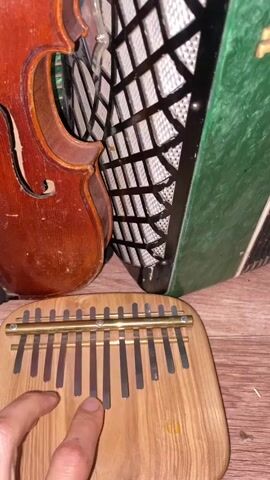 A close-up shot shows a hand playing a kalimba in front of a violin and an accordion. The kalimba is made of wood with metal tines. The violin is made of wood and has a reddish-brown color. The accordion is green and black with white bellows. The hand is pressing down on the tines of the kalimba, producing music. The background is a wooden surface.