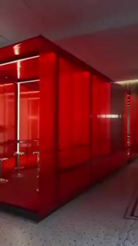 The video shows the interior of a red, cube-like structure with a minimalist design. The cube is placed within a larger, brighter space with a tiled floor. Inside the cube, there are several gold-colored stools around tables or counters. The cube is illuminated with red light throughout, accentuated by vertical and horizontal white neon lights. The walls appear to be made of translucent red panels or screens. There are no people visible inside the cube. Clothing is visible on the wall behind the structure.