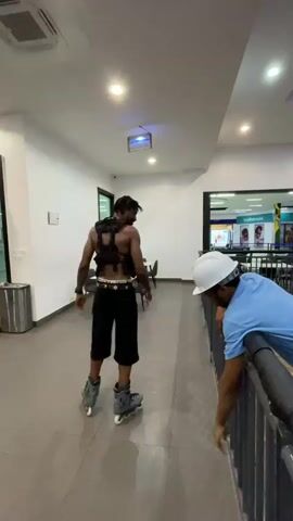 A person on roller skates wearing a black vest and black shorts performs a dance move. The person is in a hallway with tiled floors and white walls. There are tables and chairs in the background, as well as a trash can. Another person wearing a blue shirt and a white helmet leans over a railing. The person on roller skates moves their arms and body in a fluid motion, with lights on the skates flashing.