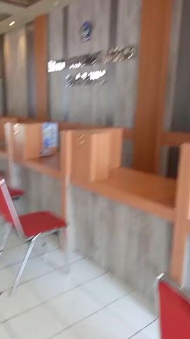 The video shows the interior of an office or service area with a reception desk. The reception desk is made of light-colored wood and has several cubicles with glass partitions. There are red chairs placed in front of the cubicles. The walls are decorated with a wood-paneled pattern. In the background, there is a logo with a blue circular emblem and some text. The video pans across the room, showing more cubicles and chairs, and then moves towards an entrance area with more seating and a sign with "BSG" and other text. The lighting is bright, and the overall impression is of a clean and organized space. There are no people visible in the immediate vicinity of the reception desk, but there is a glimpse of a person in the distance in one of the later frames.