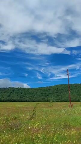 The video shows a landscape with a blue sky, white clouds, a green hill covered in trees, and a grassy field. A single, tall, rusty metal pole with wires stretches across the frame, and there are a few small trees in the field. The camera is moving, as indicated by the slight blur and the changing perspective. The overall impression is one of a rural, open space.