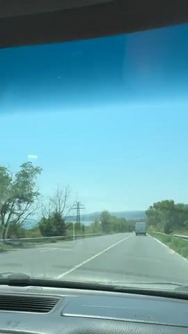 The video is filmed from the perspective of someone in a car, looking out through the windshield. The view is of a highway with two lanes. There are trees and vegetation on either side of the road. In the distance, there are rolling hills and a body of water. The sky is clear and blue. Several vehicles are visible on the road, including a white van, a dark colored SUV, and a few smaller cars. There are also power lines and utility poles visible. The sun appears to be shining brightly, creating a glare on the windshield. The car appears to be in motion.