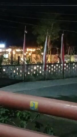 A nighttime video begins with a view of a street with a blurred orange railing in the foreground. In the background, there is a dimly lit building with string lights, and several flags with red and white horizontal stripes are visible. The video then pans to the right, revealing more of the railing and some greenery. It continues to pan, showing a yellow and blue structure with decorative patterns and text, and then a closer view of the same structure with red tubular elements in front of it. The final frames show the yellow structure in more detail, with a blue border and a sign attached to its side. There are no people visible in the video.