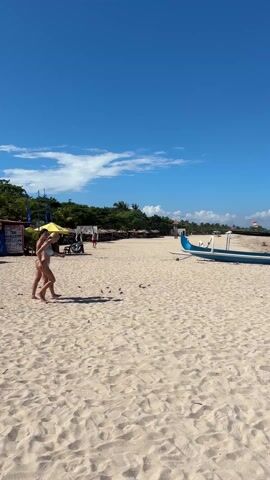 Two women in bikinis walk across a sandy beach on a sunny day. The sky is blue with scattered white clouds. A blue and white boat sits on the sand, flying a small orange flag. In the background, there is a strip of green vegetation and some beachside structures, including a yellow umbrella. A child is walking in the distance.