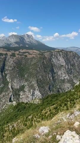 This video captures a sweeping vista of a rugged mountain landscape under a clear blue sky dotted with fluffy white clouds. The foreground features dry, yellowed grass and scattered white rocks, suggesting a high-altitude, possibly arid environment. Descending into the middle ground, the terrain becomes dramatically steep, revealing a deep canyon carved into the mountainside. The canyon walls are a mix of grey rock and dense green foliage, with patches of trees clinging precariously to the slopes. A striking turquoise lake or river is visible at the bottom of the canyon, reflecting the sunlight. In the background, a range of majestic mountains stretches across the horizon, their peaks softened by distance. The overall impression is one of immense natural beauty and scale, with the raw power of the earth evident in the dramatic topography. There are no people or man-made objects visible in the frame. The camera slowly pans across the scene, allowing for a thorough appreciation of the vastness and ruggedness of the landscape.