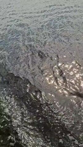 This video shows a wide body of water, likely a river, with a shoreline visible in the distance. The water's surface is rippled, reflecting the sunlight in shimmering patches. The shoreline is lined with trees and dotted with houses, suggesting a rural or semi-rural setting. A hill rises behind the shoreline, with buildings and communication towers visible on its crest. The sky is partly cloudy, with patches of blue sky showing through white clouds. The overall impression is of a peaceful, sunny day in a scenic landscape. The timestamp indicates this footage was captured on July 26, 2023, at an unknown time of day, likely daytime given the sunlight. The location is Verhnij Uslon, Russia.