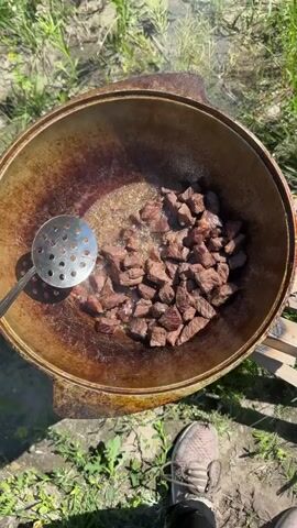 A person is cooking diced meat in a large, round, cast-iron pot over an open fire. The pot is dark brown and appears well-used. The meat is in bite-sized pieces and is browning in hot oil. A slotted spoon is used to stir the meat. The scene appears to be outdoors, with green grass and dirt visible around the pot. The person's legs and feet, wearing athletic shoes, are visible at the bottom of the frame. The lighting suggests it is daytime.