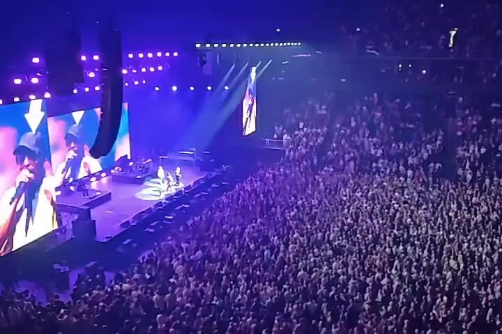 An aerial view shows a large, packed concert venue with a stage set up on the left side of the frame. A massive screen behind the stage displays a close-up of a person wearing a hat and holding a microphone. Below the screen, on the stage, several musicians are performing, with bright purple and white lights illuminating the scene. The audience, a dense mass of people, fills the floor and stadium seating on the right, their faces turned towards the stage. The overall atmosphere is energetic and lively, indicative of a live musical performance.