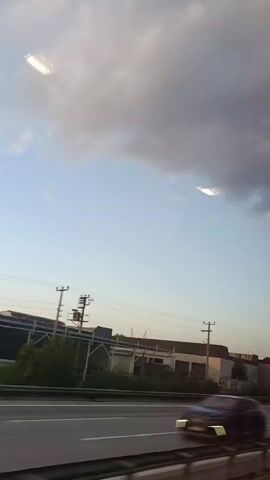 This video is an exterior shot of a highway scene taken from a moving vehicle. In the foreground, there is a highway with multiple lanes of traffic. A white car is visible driving in the right lane. In the background, there are industrial buildings, power lines, and trees. The sky is partly cloudy with a mix of blue sky and dark clouds. Two bright, rectangular objects are visible in the clouds.
