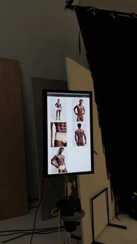 A monitor displays a grid of six photographs of a male model in underwear, shown from various angles. The monitor is mounted on a stand that also holds a camera. The scene appears to be a photography studio, with large white and dark panels in the background.