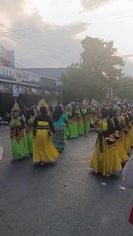 A group of dancers in brightly colored costumes, predominantly yellow and green, are performing in a street parade. The dancers are arranged in rows and appear to be moving in unison. Some are holding small objects, possibly instruments or props. The background shows a street lined with buildings, including a shop with signage that reads "TOKO DUTA". Power lines and trees are visible in the distance. The sky is cloudy, with sunlight breaking through. The overall atmosphere suggests a cultural event or celebration.