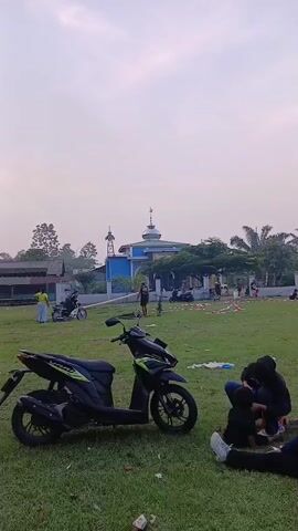 A black and green scooter is parked on a grassy field. A group of people are sitting on the grass in the foreground, some are interacting with each other. In the background, there is a blue and white building with a dome, which appears to be a mosque. Other people are visible in the middle ground, some are standing or walking, and there are a few motorcycles parked. The sky is overcast and the lighting suggests it is either early morning or late afternoon. The field appears to be a public space, possibly a park or a community area. There is some scattered debris on the grass.