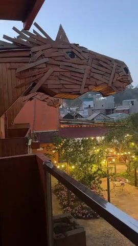 The video presents a scenic view of a courtyard at dusk, possibly in a Southeast Asian location. The foreground is dominated by a large, wooden sculpture resembling a horse's head, constructed from numerous rectangular planks. The camera is positioned on a balcony with a glass and metal railing. Below, the courtyard is illuminated by strings of warm, yellow lights strung between trees, creating a festive atmosphere. A stone pathway meanders through the garden, leading to a small seating area with a table and chairs. Beyond the courtyard, a collection of buildings with terracotta tile roofs can be seen, giving context to the location being likely an urban or suburban setting. The sky is a soft, muted blue, indicating either early evening or late afternoon. There is no activity taking place, and no people are visible in the shot.