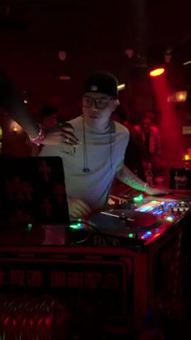 A DJ stands behind a turntable setup in a dimly lit club. The DJ, wearing a light blue t-shirt, glasses, and a baseball cap, is focused on the equipment. A laptop with a cross design is positioned to the left of the DJ. In the background, two other individuals are visible, one in a plaid shirt sitting down, and another standing. The environment is characterized by changing colored lights, including red, green, and blue. There are also decorative brick walls and lights in the background.