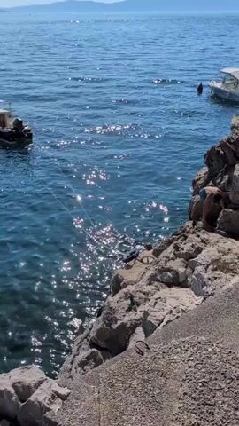 A person dressed in blue shorts and a light-colored top stands on a rocky cliff overlooking the sea. The water is a deep blue and sparkles in the sunlight. A white boat is visible in the distance. The person appears to be preparing to jump or dive into the water.