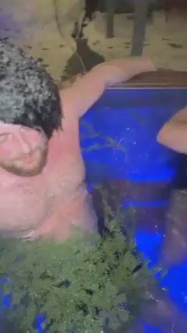 Two shirtless men are submerged in a container filled with water illuminated by blue lights. The container appears to be made of wood. Greenery is placed around the edge. One man is partially visible and moves his arms in the water. The camera moves, showing the men submerged up to their necks.