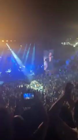 A crowded concert venue is shown in the video. A lit stage is visible in the distance with performers on it, illuminated by bright spotlights. There's a large screen showing the performer behind the stage. The audience is densely packed, many holding up their phones to record the performance. In the foreground, people’s arms are raised in the air. The general atmosphere appears energetic.