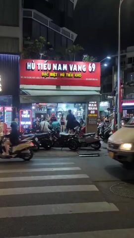 The video shows the exterior of a restaurant at night. The restaurant's name is "Hủ Tiếu Nam Vang 69" with a large sign in red with white letters. The sign indicates it specializes in "Hủ Tiếu Khô," which translates to dry rice noodles. People are seated and eating inside the restaurant, visible through the glass front. Several motorcycles are parked in front. A white van passes in the foreground. The address on the sign is "69 Trần Huy Liệu, P. 12, Q. Phú Nhuận," a location in Ho Chi Minh City, Vietnam. The phone number "0937 276 744" is also displayed on the sign. Motorbike traffic continues to move in the background.
