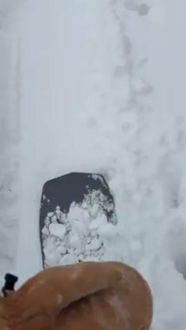 A person is snowboarding in a snowy environment. The camera is positioned close to the snowboard, providing a view of the board and the snow beneath it. The snowboard is gray and covered in clumps of snow. The person is wearing blue snowboarding pants. The snow appears to be deep, creating tracks in the snow as the snowboard moves. The overall scene conveys a sense of winter activity and movement through snowy terrain.