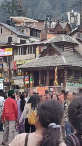 The video captures a bustling street scene, likely in a mountainous region, filled with a diverse crowd of people. The architecture suggests a blend of traditional and modern influences, with multi-story buildings adorned with various signs and decorations. The shops on either side of the street are packed with customers, creating a lively atmosphere. Some individuals are seen carrying backpacks, indicating they might be tourists. The presence of shops, restaurants, and the general ambiance suggest a popular tourist destination.
