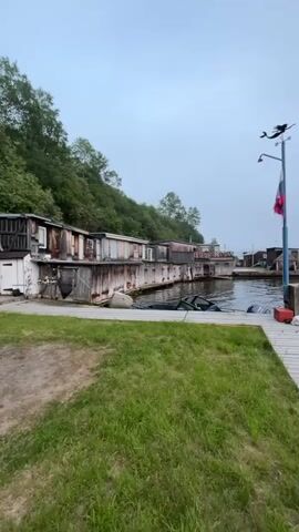 This media file depicts a waterfront scene in Sovetskaya Gavan', Russia, likely during the early morning or late evening due to the overcast sky and muted lighting. The overall mood is quiet and slightly melancholic, enhanced by the fog rolling in from the sea.

The primary subjects are a series of weathered, rustic structures situated on a dock or pier. These buildings appear to be small homes or workshops, characterized by their aged wooden siding and simple construction. Some have visible windows, and one structure has what looks like a boat or some debris partially submerged in the water next to it.

In the foreground, a well-worn wooden boardwalk extends into the water, with a patch of green grass to its left. On the boardwalk, a tall metal pole stands, adorned with a weathervane shaped like an airplane and a Russian flag, which is partially unfurled. Scattered around the base of the pole are a few objects, including what appear to be orange buoys and some white bags.

The background reveals a body of water, likely a bay or harbor, with more distant structures visible on the opposite shore, shrouded in mist. A green, tree-covered hill rises behind the buildings on the left side of the frame. The weather is clearly foggy, obscuring the horizon and lending a sense of remoteness and isolation to the scene.

There are no visible people in the media. The activity seems to be centered around the quiet existence of this waterside community. The time of day is suggested to be either dawn or dusk by the soft, diffused light and the pervasive fog. The location cues (Russian flag, the name Sovetskaya Gavan' provided) firmly place the scene in Russia. The overall impression is one of a remote, hardworking coastal settlement experiencing a typical, perhaps damp and misty, day. No discernible text is visible within the frame.