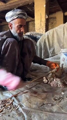 An older man with a beard and a white and black turban is squatting on a clear plastic sheet. He is wearing a brown outfit and black boots. He has orange gloves on one hand and a pink glove on the other. He appears to be cleaning a pile of chicken organs. There are many long, tube-like organs and a pile of fleshy organs on the plastic. In the background, there is a brick wall and a wooden structure. There is a white bucket or container to the right of the man. Some brown wool is piled behind him. The environment appears to be a barn or storage area.