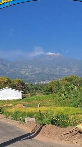 A landscape video shows a mountain range in the background under a clear blue sky with a few wispy clouds. In the midground, there is a white building with a dark roof and terraced fields of green and yellow crops. A person in white clothing is visible working in one of the fields. A dirt road with yellow caution tape runs along the bottom of the frame. The top of the frame is cut off by what appears to be the bottom of an archway with some text on it.