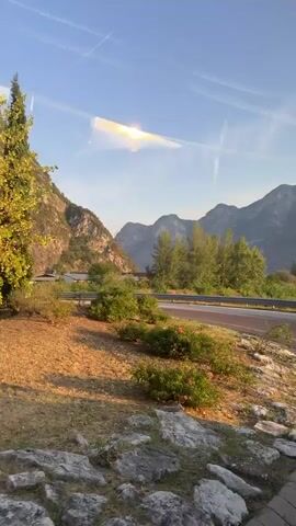 This is a vertical, point-of-view shot taken from a moving vehicle, likely a car or bus, looking out at a mountainous landscape. The time appears to be late afternoon or early evening, indicated by the warm, golden light illuminating the rock faces of the mountains and the clear blue sky.

In the foreground, to the left and center, is a grassy embankment dotted with rocks and low-lying shrubs. A tall, slender cypress tree stands prominently on this embankment. To the right, a curved road, possibly a highway or a scenic route, is visible. The road has a red lane marker. The road curves out of frame to the right.

Behind the embankment and road, the majestic mountains dominate the scene. They are characterized by steep, rocky slopes, with visible strata and some areas of lighter colored rock. Patches of vegetation cling to the mountainsides. A large, dark opening in the mountain, appearing to be a tunnel entrance, is visible further back on the right side of the frame. Road signs are also visible near the tunnel entrance.

The lighting creates a sense of depth and highlights the textures of the mountains and the surrounding vegetation. The overall impression is one of natural beauty and passage through a mountainous region. There are no people or animals visible in the frame. The movement of the camera suggests a journey is in progress.