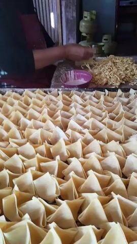 A close-up shot shows rows of uncooked dumplings arranged neatly on a tray. In the background, a person wearing an apron is seen preparing more dumplings. A pile of noodles and a bowl with some white liquid are also visible. The scene appears to be in a kitchen or food preparation area.