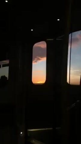 The video shows a view from inside a train car. It is dark inside the train. Through a window, a sunset is visible, with orange and pink clouds. Another window shows more of the sunset and a silhouette of trees.