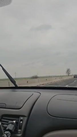 The video shows a car ride on a highway during a cloudy day. The view is from the driver's seat, focused on the road ahead.  Wipers are actively cleaning the windshield.  The road has concrete barriers and a white lane marker.  The landscape outside consists of flat fields with power lines and the occasional tree.  There is another car visible in the distance. Loud music is playing in the car.