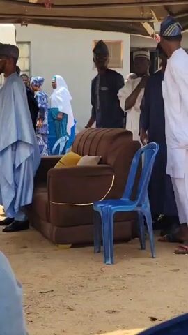 Here's a detailed description of the video:

The video shows a gathering of people outdoors, possibly at a social event or ceremony. Several men dressed in traditional African attire are standing and talking. One man is wearing a light blue robe-like garment, another is in white, and others are in darker-colored outfits with patterned hats.

In the background, more people can be seen, including women wearing headscarves and colorful dresses. Some individuals are seated, while others stand or move around. A brown sofa with gold trim and a couple of blue plastic chairs are placed on the ground. The setting appears to be a yard or open space, possibly near a building. 

The video seems to capture a candid moment of people socializing and interacting with each other.