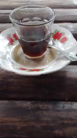 A close-up shot shows a small glass of dark liquid, likely coffee, sitting on a saucer with a floral design. A silver spoon rests on the saucer next to the glass. The glass and saucer are on a surface made of dark, horizontal wooden planks, which appear to be part of a table or bench. In the background, out of focus, is a blurry image of a motorcycle. The lighting suggests an outdoor setting.