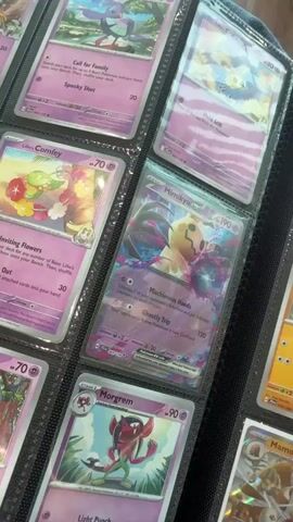 The video shows a close-up view of a binder filled with Pokémon cards. The cards are displayed in plastic sleeves, and the binder is open, revealing multiple pages of cards. The focus is on a particular card featuring the Pokémon Mimikyu, which is shown in the center of the frame. Other Pokémon cards are visible around it, with varying artwork and text. The lighting suggests the video was filmed indoors, possibly in a room with ambient light. The camera appears to be held by hand, as there is slight movement and the focus shifts occasionally. The overall scene is a collection of trading cards.