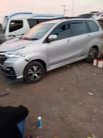 A silver minivan is parked on a dirt lot. The front right fender of the minivan is damaged, appearing dented and scraped. A white truck is parked behind the minivan. A person is sitting on the ground in the foreground, facing away from the camera. A bottle of water is on the ground near the person. The sky in the background is a pale pink and orange, suggesting it is either sunrise or sunset. The rear hatch of the minivan is open, and two brown bags are visible inside.