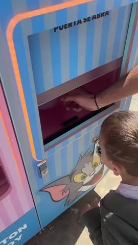 A man and child stand in front of a cotton candy vending machine that has the cartoon character Tom from Tom and Jerry on its exterior. The man is retrieving cotton candy from the machine, and hands it to the child.