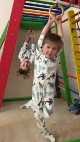 Two young children, both wearing patterned pajama suits, play on an indoor climbing structure. The structure consists of colorful bars and a net. One child hangs upside down from rings, while the other stands and holds onto a ring. The background includes carpeted flooring and a partially visible wall.