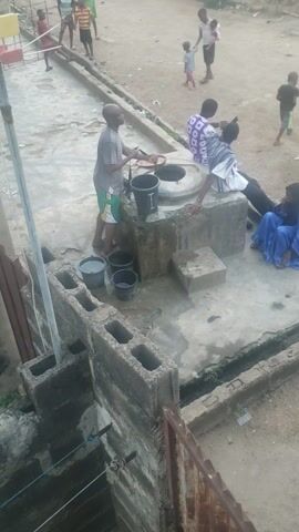 The video captures a daytime community scene in Alalubosa, Nigeria, primarily focused on a communal water well. Several people are gathered around the well area. A man is seen actively drawing water from the well using a rope and a black bucket. Other black and grey buckets are placed on the concrete ground nearby, indicating more water collection. A woman dressed in a blue garment is sitting next to the well structure, alongside another person. Children are visible in the background, moving and playing on the dirt ground.

The camera pans to reveal more of the surroundings, showing a mix of dirt ground and concrete paths. A drainage ditch or trench runs alongside the main activity area, containing some scattered debris and trash. In the background, simple residential buildings with corrugated metal structures are visible, and a motorcycle is parked. A tall concrete pole stands prominently. The overall setting suggests a residential neighborhood where the well serves as a vital water source. The weather appears clear, and people are engaged in everyday activities, primarily water fetching and socializing. One of the black buckets has "FARMERS" printed on its side in white letters.