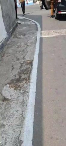 The video begins with a close-up view of a cracked concrete sidewalk next to a wall. The scene then pans out to reveal several people standing on a paved road, some near the sidewalk and others further down the road. Indonesian flags are visible in the background, and there are trees and buildings. A black car is parked on the side of the road, and its rear is shown with a sticker displaying text. Another person in a camouflage uniform with a red box on their head walks past the other individuals on the road. The video appears to capture a moment in a public outdoor setting.